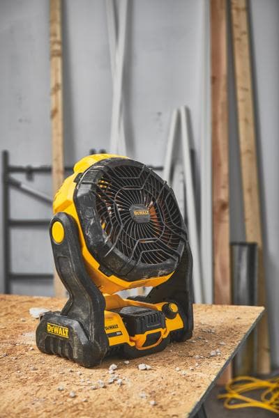 A DEWALT portable cordless fan (model DCE512B) is placed on a wooden workbench in a workshop with construction materials in the background.