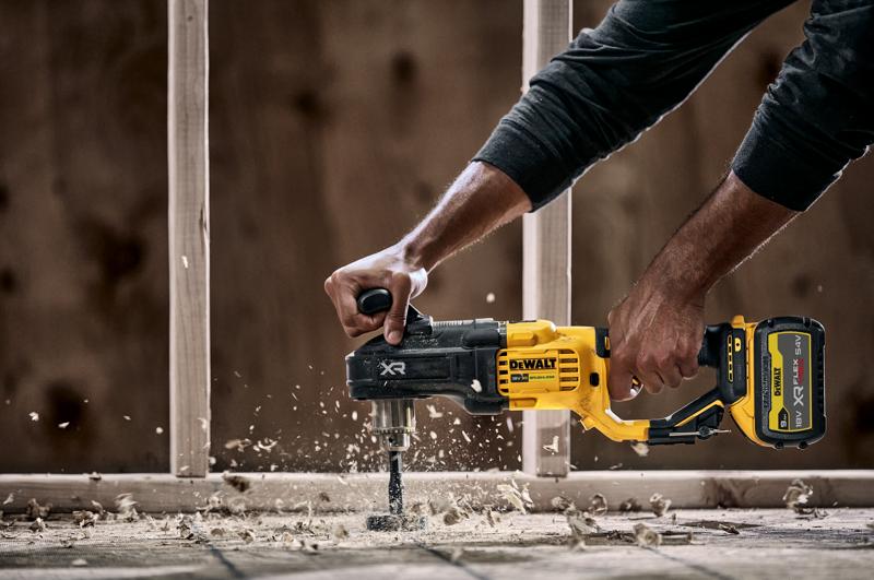 A person using a DEWALT DCD444N cordless power drill with a side handle to drill through wood, creating wood shavings and dust. Construction framing is visible in the background.