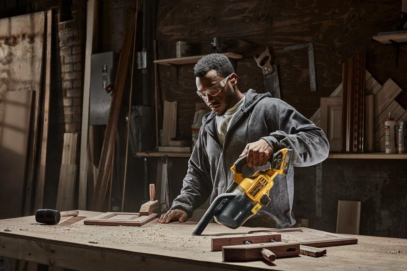 A person using a DEWALT portable vacuum cleaner model DCV501HB to clean sawdust from a workbench in a woodworking shop. Various tools and wooden pieces are visible on the table and shelves in the background.